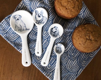 May include: Set of four white ceramic measuring spoons with dog illustrations. The spoons are labeled 1/4 teaspoon, 1/2 teaspoon, 1 teaspoon, and 1 tablespoon. The spoons are on a blue and white patterned fabric with two brown muffins.