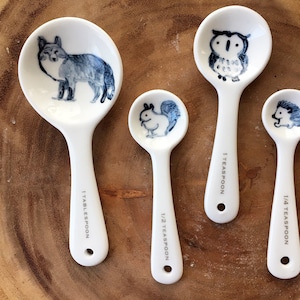 May include: Set of four white ceramic measuring spoons with blue and white animal illustrations. The spoons are labelled "1 Tablespoon", "1 Teaspoon", "1/2 Teaspoon", and "1/4 Teaspoon".