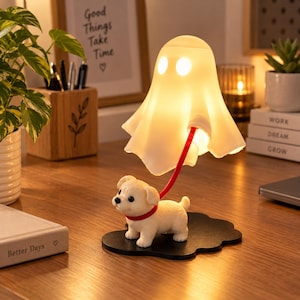 May include: A white ghost-shaped lamp with two circular cutouts for eyes, illuminated from within. The ghost is attached to a red leash held by a small, cream-colored puppy figurine standing on a black, cloud-shaped base on a wooden surface.