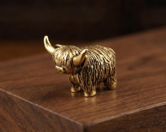 May include: A small, solid brass figurine of a Highland cow. The metal sculpture features a textured, shaggy coat, curved horns, and a detailed face, capturing the distinct appearance of the animal in a compact, metallic form.