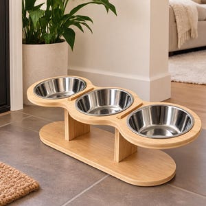 May include: A triple pet feeder station featuring three removable stainless steel bowls set into a light-toned wooden stand. The stand has a smooth, oval-shaped base and two vertical supports that hold the elevated platform for the bowls.