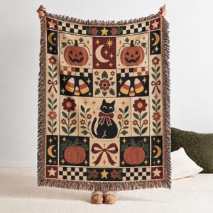 Könnte beinhalten: Eine gewebte Wohndecke mit einem Gittermuster, das Illustrationen von Kürbissen, einer schwarzen Katze mit roter Schleife, Zuckermais, Mondsicheln, Sternen, Blumen und Schleifen in den Farben Orange, Schwarz, Creme und Rot zeigt.