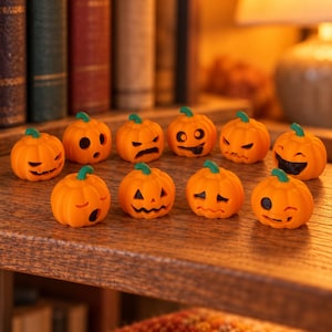 May include: A collection of ten small orange pumpkin figurines, each featuring a unique carved facial expression with black detailing. The miniature pumpkins have green stems and are arranged in two rows on a textured wooden surface.