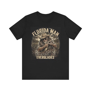 May include: A black short-sleeve t-shirt featuring a graphic print of a person wearing a cowboy hat and sunglasses while holding a large snake. The text FLORIDA MAN is arched above the image, and the word EVERGLADES is printed below the graphic.