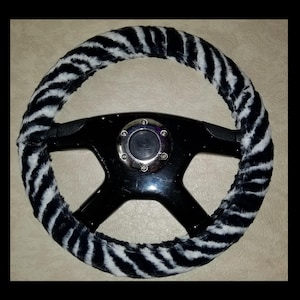 May include: A black steering wheel with a zebra-striped, plush cover. The cover is black and white, with a soft, furry texture. The center of the wheel has a chrome emblem.