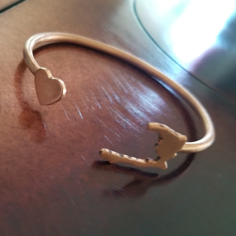May include: A gold-toned bracelet with a heart and a state outline charm. The bracelet is on a wooden surface.