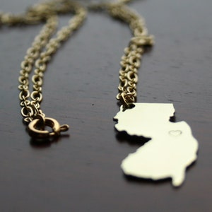 Può includere: Collana color oro con un ciondolo a forma dello stato del New Jersey. Il ciondolo presenta un piccolo dettaglio a forma di cuore. La catena è un design delicato e collegato con chiusura. La collana è mostrata su una superficie marrone scuro.