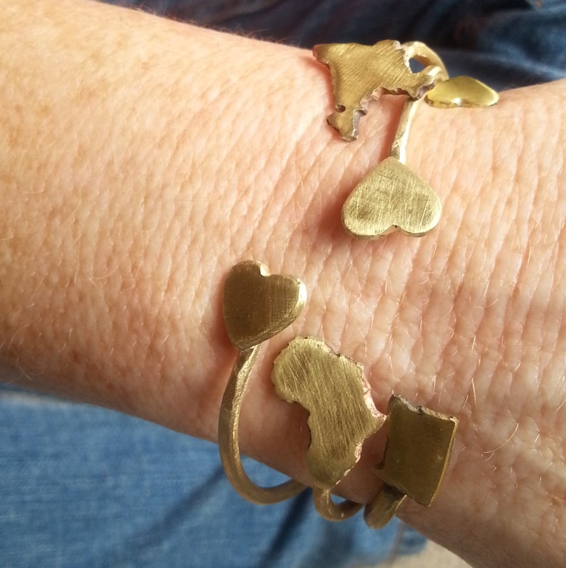 May include: A gold bracelet with a heart and a map of Africa design. The bracelet is on a person's wrist.
