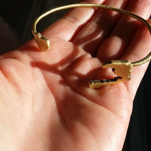 May include: A gold-toned metal bracelet with a unique, open-ended design. The bracelet is made of a single piece of metal that is bent into a circle and then has two open ends that are shaped like jagged edges.