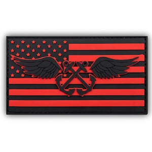 May include: A rectangular PVC morale patch featuring a stylised American flag design in black and red. The design includes a central emblem of two crossed anchors with large black wings extending outward, set against a field of red stars and stripes.