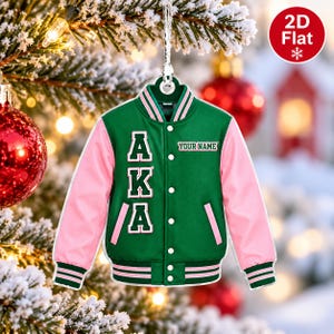 May include: A 2D flat Christmas tree ornament shaped like a green and pink varsity jacket. The jacket features the letters AKA in white with green outlines, the text YOUR NAME, white snap buttons, and striped ribbed trim on the cuffs and hem.