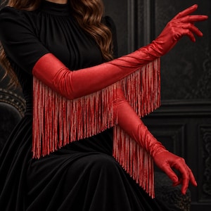 May include: A pair of vibrant red opera-length gloves featuring long, flowing fringe detailing that extends from the forearm down to the wrist. The gloves are crafted from a smooth, satin-like material and reach above the elbow, providing a bold look.