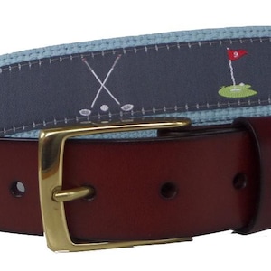 Puede incluir: Un cinturón de cuero marrón con una hebilla dorada. La sección superior del cinturón presenta una tela azul con un patrón temático de golf, que incluye palos de golf, pelotas de golf y banderas. El cinturón está diseñado para uso casual.