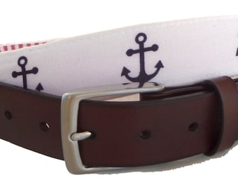 May include: A brown leather belt with a silver buckle. The white fabric belt features navy blue anchor designs. The interior lining has a red and white striped pattern. The belt is a nautical-themed accessory.