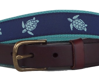 May include: A brown leather belt with a brass buckle. The belt features a navy blue fabric band with light blue turtle designs. The belt is a stylish accessory for casual or formal wear.