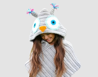 May include: A grey crocheted owl poncho with a blue and white eye and an orange beak. The poncho has a hood with a pink and blue pom-pom on top.