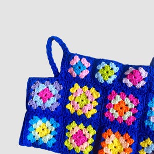 May include: A blue crochet top with colorful granny squares. The top has two straps and is made with a variety of bright colors, including pink, yellow, green, and orange.