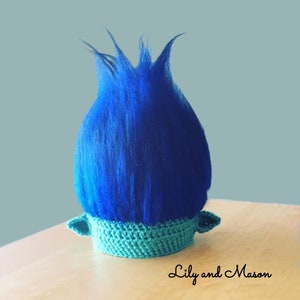 May include: Blue and teal crocheted troll hat with blue yarn hair. The hat has two small ears on the sides.