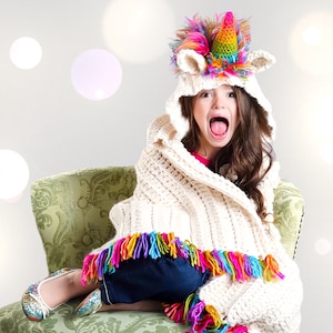May include: A young girl wearing a white crocheted hooded blanket with colorful fringe and a unicorn head hat. She is sitting on a green patterned chair with her mouth open and tongue sticking out.