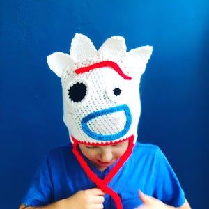 May include: A white crocheted hat with a red and blue face design, resembling a popular cartoon character. The hat has long ties that can be tied under the chin.