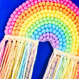 May include: A rainbow-colored crocheted wall hanging with long, colorful tassels. The rainbow is made of small, textured balls of yarn.