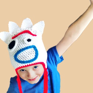 May include: A child wearing a white crocheted hat with a red, blue, and black cartoon face. The hat has long red ties.