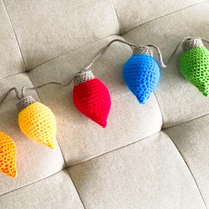 May include: A string of crocheted light bulbs in various colors, including red, orange, yellow, green, and blue. The bulbs are connected by a gray string and are arranged in a row on a light gray surface.