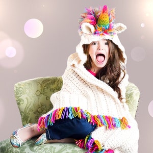 May include: A young girl wearing a white crocheted blanket with colorful fringe sits in a green chair. She is wearing a unicorn hooded hat with a rainbow horn and a pink and white mane. The girl is smiling and looking at the camera.