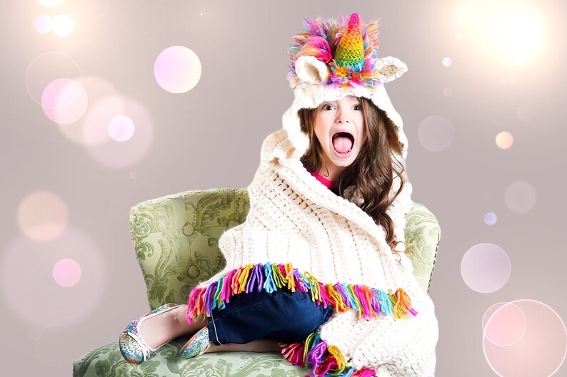 May include: A young girl wearing a white crocheted blanket with colorful fringe sits in a green chair. She is wearing a unicorn hooded hat with a rainbow horn and a pink and white mane. The girl is smiling and looking at the camera.