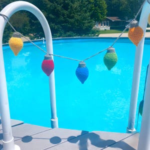 May include: A string of crocheted lightbulb ornaments in various colors hangs over a blue swimming pool. The ornaments are yellow, red, blue, green, and orange.