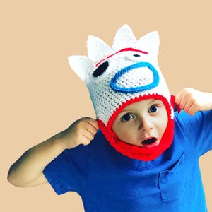 May include: A white crocheted hat with a red and blue face design. The hat has a red brim and a white pom-pom on top. The hat is being worn by a child.