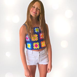 May include: A young person wearing a blue crochet top with colorful squares and white denim shorts.