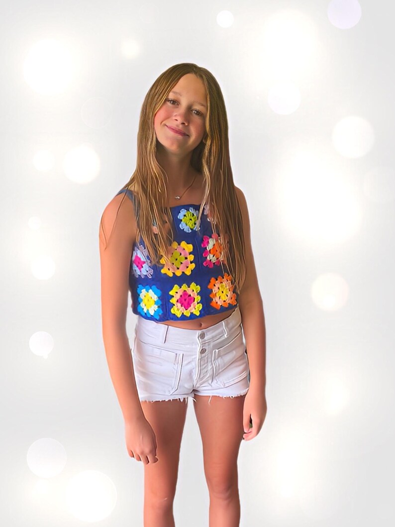 May include: A young person wearing a blue crochet top with colorful squares and white denim shorts.