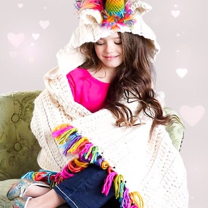 May include: A young girl wearing a white hooded blanket with a rainbow unicorn horn on top. The blanket has a colorful fringe trim. She is sitting on a green chair.