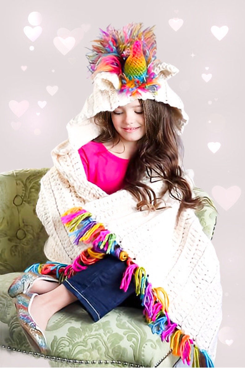 May include: A young girl wearing a white hooded blanket with a rainbow unicorn horn on top. The blanket has a colorful fringe trim. She is sitting on a green chair.