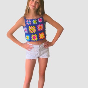May include: A young person wearing a blue crochet top with colorful squares and white denim shorts. They are wearing white socks and have their hands on their hips.