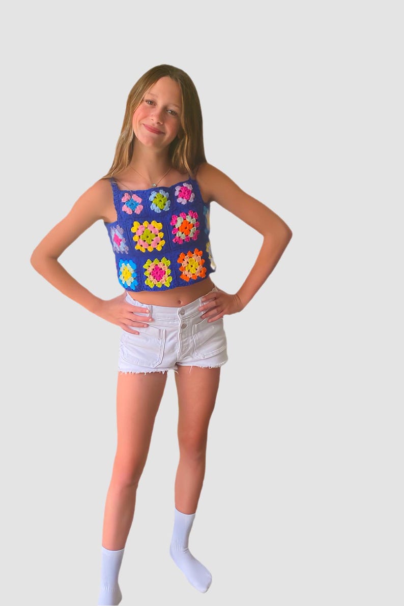 May include: A young person wearing a blue crochet top with colorful squares and white denim shorts. They are wearing white socks and have their hands on their hips.