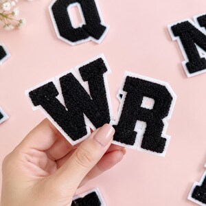 May include: Black varsity-style letter patches with a textured, fuzzy surface and a white border. The letters W and R are held together, while other individual letter patches like Q, J, and N are scattered around on a light pink background.