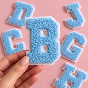 May include: A light blue letter B patch with a textured, fuzzy surface and a white border. The letter features a block-style font with a cutout center. Other similar light blue letter patches are visible in the background.