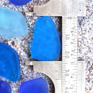 May include: Close-up of several blue sea glass pieces with a metal ruler measuring one piece. The sea glass is various shades of blue, with one piece being measured at approximately 1.5 inches. The sea glass is on a sandy surface.