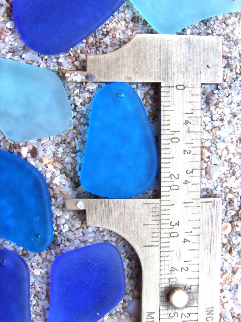 May include: Close-up of several blue sea glass pieces with a metal ruler measuring one piece. The sea glass is various shades of blue, with one piece being measured at approximately 1.5 inches. The sea glass is on a sandy surface.