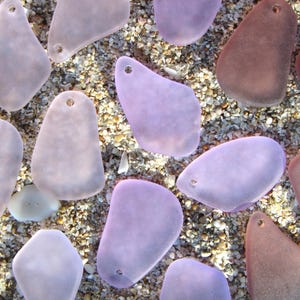 May include: An overhead shot of sea glass pendants in various shades of purple and brown, each with a small hole for stringing. The pendants are scattered on a bed of sand and small pebbles, creating a natural, beachy aesthetic.
