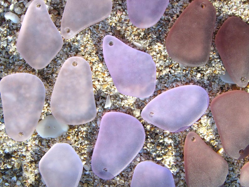 May include: An overhead shot of sea glass pendants in various shades of purple and brown, each with a small hole for stringing. The pendants are scattered on a bed of sand and small pebbles, creating a natural, beachy aesthetic.