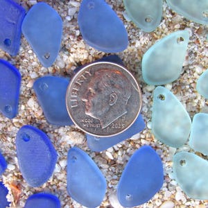 May include: A collection of small, frosted sea glass pieces in shades of deep blue and pale aqua, each featuring a single drilled hole. A silver-colored United States dime coin is placed in the center for scale, showing the size of the glass pieces.