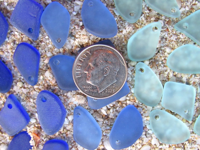 May include: A collection of small, frosted sea glass pieces in shades of deep blue and pale aqua, each featuring a single drilled hole. A silver-colored United States dime coin is placed in the center for scale, showing the size of the glass pieces.