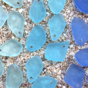 May include: A collection of frosted sea glass pendants in various shades of light blue and deep cobalt blue. Each piece features a smooth, matte texture and a single pre-drilled hole at the top, suitable for jewelry making and craft projects.