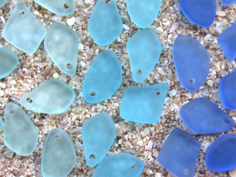 May include: A collection of frosted sea glass pendants in various shades of light blue and deep cobalt blue. Each piece features a smooth, matte texture and a single pre-drilled hole at the top, suitable for jewelry making and craft projects.