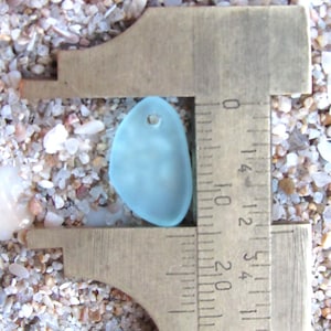 May include: A smooth, frosted light blue sea glass pendant with a single drilled hole at the top. The piece is positioned within a brass caliper, showing a width of approximately 1.5 centimeters on the metric scale of the measuring tool.