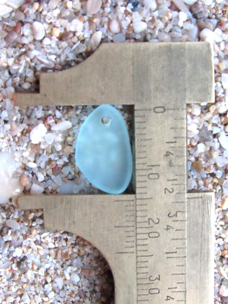 May include: A smooth, frosted light blue sea glass pendant with a single drilled hole at the top. The piece is positioned within a brass caliper, showing a width of approximately 1.5 centimeters on the metric scale of the measuring tool.