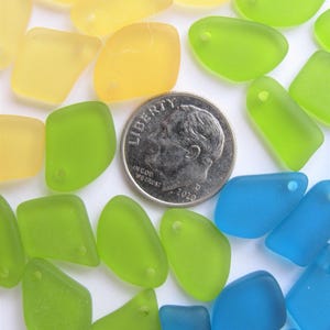 May include: A collection of translucent, frosted glass pieces in yellow, lime green, and blue, each featuring a small pre-drilled hole for jewelry making. A silver-colored United States dime is positioned among the pieces to provide a scale reference.